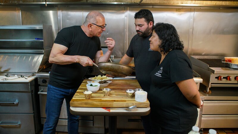 Chef Robert Irvine shows Omar Barreiro, owner and chef of Amistad Grill, how to make the entrées from the new menu at Amistad Grill in Homestead, FL; as seen on Food Network’s Restaurant: Impossible Season 20. – Bild: Warner Bros. Discovery