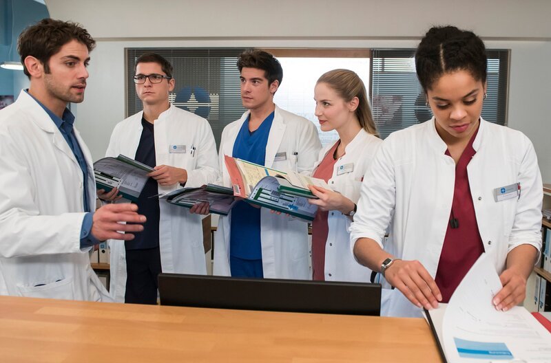 Als Übungsaufgabe verteilt Niklas (Roy Peter Link, l.) alte Patientenfälle, welche die Assistenzärzte Elias (Stefan Ruppe, 2.v.l.), Ben (Philipp Danne, 3.v.l.), Julia (Mirka Pigulla, 2.v.r.) und Vivi (Jane Chirwa, r.) bis zum nächsten Tag bearbeiten müssen. – Bild: ARD/​Jens Ulrich Koch