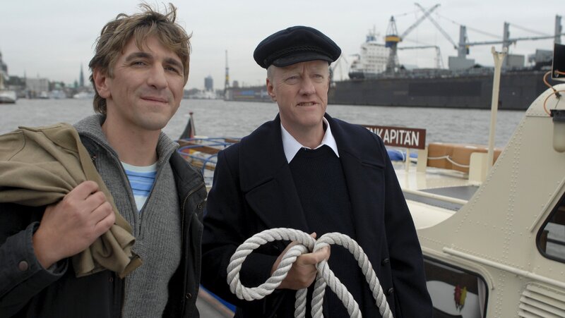 Fritz Fuchs (Guido Hammesfahr, l.) will an Bord der Cap San Diego, doch wo findet er sein Schiff? Der Barkassenkapitän (Axel Olsson, r.) nimmt Fritz mit auf eine Fahrt durch den Hafen. – Bild: ZDF