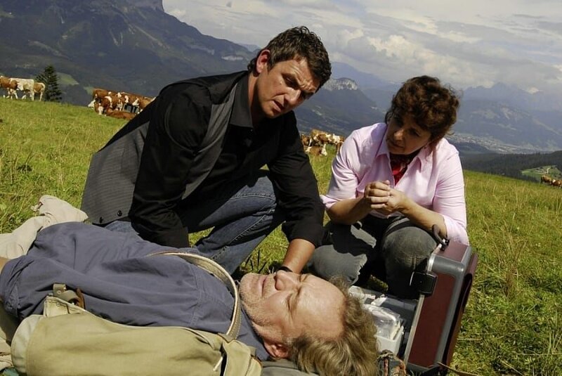 Dr. Martin Gruber (Hans Sigl, l.) und seine Mutter Lisbeth (Monika Baumgartner, r.) kümmern sich um den kollabierten Julius (Gerd Silberbauer, vorne). – Bild: port.hu