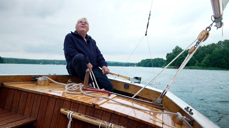 Wenige Kilometer südöstlich von Berlin liegt der Scharmützelsee, das „Märkische Meer“, mit traumhaftem Sandstrand, glitzernden Booten und Künstlerinnen und Künstlern, die hier mitreißende Musik, mehrstöckige Torten und das idyllischste Filmfestival Brandenburgs machen. – Klaus Rattemayer segelt auf der Oceania. – Bild: rbb/​Dennis Wagner