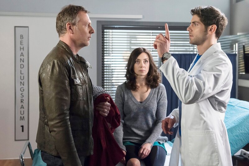 ARD PROGRAMMDIREKTION, IN ALLER FREUNDSCHAFT – DIE JUNGEN ÄRZTE, neue Vorabend-Weekly, Folge 21  „Schritt für Schritt“, am Donnerstag (09.07.15) um 18:50 Uhr im Ersten. Eigentlich lässt sich Susanne Albrecht (Julia Richter, M.) von Dr. Ahrend (Roy Peter Link, r.) untersuchen, doch weil sich ihr Mann Jochen Hoffmann (Christoph Grunert, l.) anscheinend etwas ungeschickt anstellt und am Bett anstösst, nimmt Dr. Ahrend auch an ihm eine kleine Untersuchung vor. Offenbar ist sein Blickfeld links eingeschränkt. – Bild: ARD/​Jens Ulrich Koch