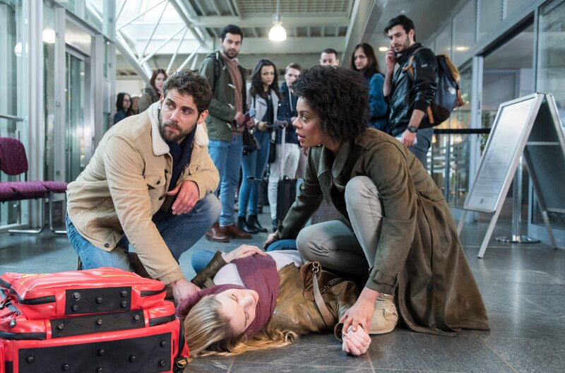 Niklas (Roy Peter Link, l.) eilt mit einem RTW sogleich zum Flughafen und trifft dort auf Vivi (Jane Chirwa, r.) und die ohnmächtige Julia (Mirka Pigulla, M. mit Komparsen h.). – Bild: ARD/​Jens Ulrich Koch