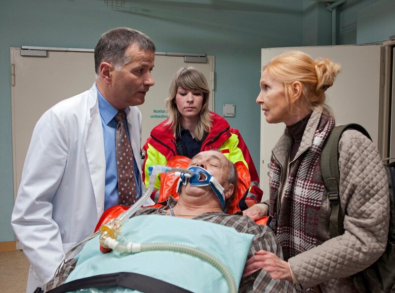 Als Prof. Simoni (Dieter Bellmann, liegend) von der Notärztin (Komparsin) und Oberschwester Ingrid (Jutta Kammann, rechts) in die Sachsenklinik eingeliefert wird, wartet Dr. Roland Heilmann (Thomas Rühmann) schon auf sie. – Bild: MDR/​MDR/​Krajewsky