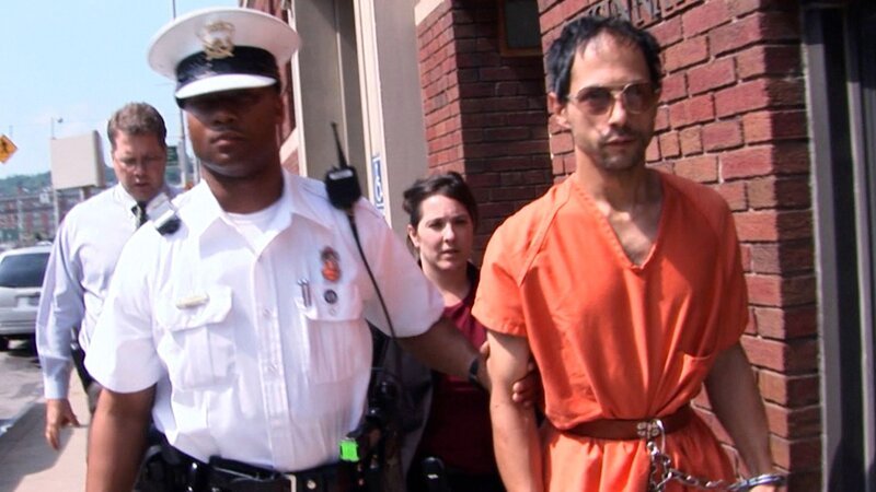 Der Polizist nimmt den Gefangenen im Beisein eines Mannes und einer Frau, die hinter ihnen gehen, in Gewahrsam – Bild: Crime + Investigation /​ A+E Networks