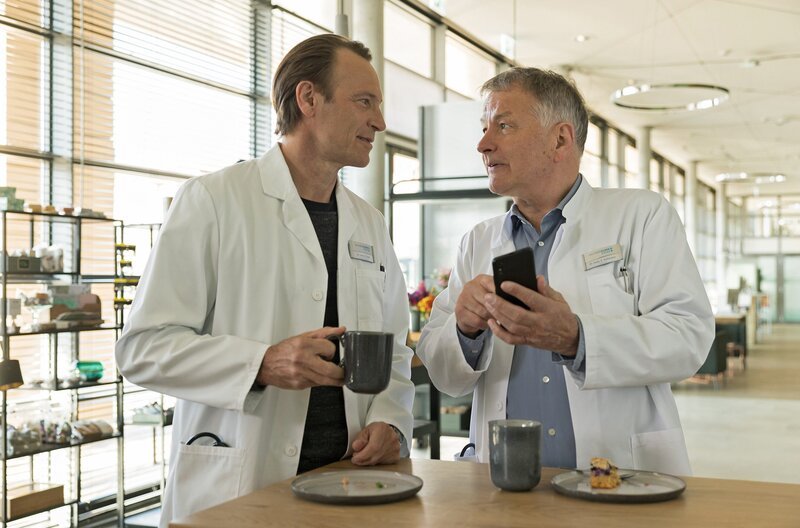Dr. Roland Heilmann (Thomas Rühmann, re.) hat ein bisschen über Darren Macneil, dem neuen Physiotherapeuten, recherchiert. Dieser ist, nach Rolands Absage, am Abend mit Kathrin Globisch verabredet. Darren ist auch Personal-Trainer und obwohl sich Dr. Martin Stein (Bernhard Bettermann, li.) ein wenig über seinen Freund lustig macht, bucht Roland eine Stunde bei Darren. – Bild: MDR/​Saxonia Media/​Rudolf Wernicke