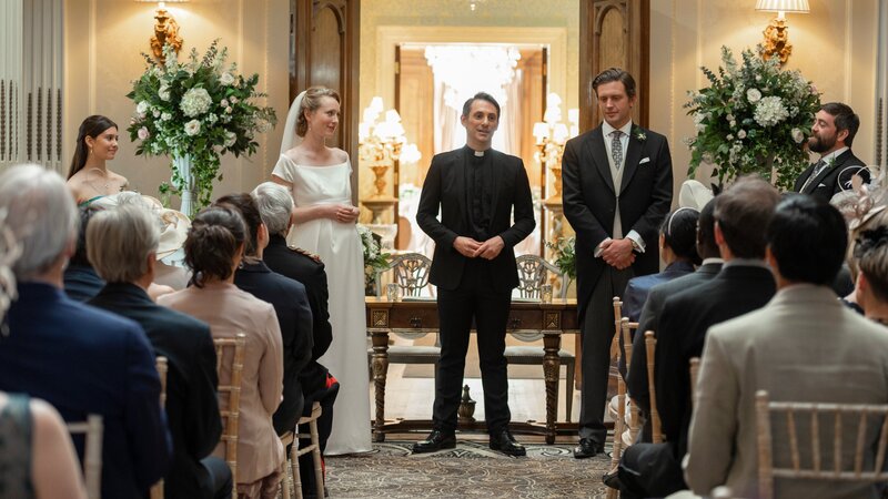 Nach langem Warten traut der Pfarrer (Ashley Cook, M.) endlich Greg (Tom Durant Pritchard, r.) und Emma (Alice Orr-Ewing, l.) zum Ehepaar. Was die beiden dabei aber noch nicht wissen, ist, dass sie ein ganz besonderes Hochzeitsgeschenk erwartet. – Bild: Anika Molnar /​ ZDF