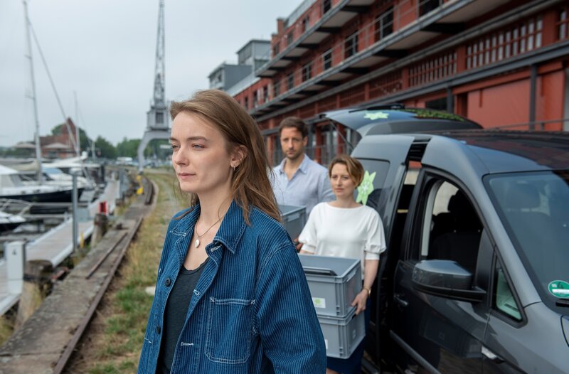 Emily (Hannah Dalmeyer, l.) blickt melancholisch auf das Boot ihrer verstorbenen Freundin. Im Hintergrund laden Marold (Dominik Maringer, M.) und Viktoria (Wanda Perdelwitz, r.) Kisten aus dem Auto. – Bild: ARD/​Thorsten Jander