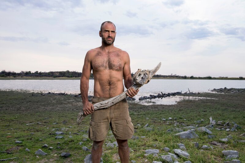 Ed Stafford holding a tool he has made in front of watering hole landscape, Namibia. – Bild: Discovery Communications