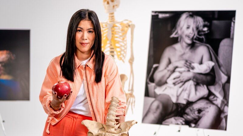 Mai Thi Nguyen-Kim im Studio, umgeben von Fotos von Neugeborenen. Mit Hilfe eines Granatapfels und eines Beckens erklärt sie den Geburtsprozess beim Menschen. – Bild: ZDF