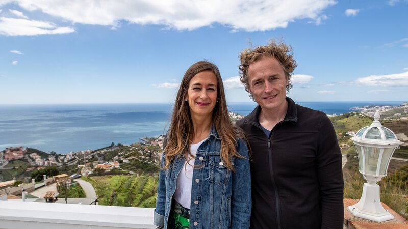 Episode 004 – Holly Prole (L) and Jason Prole (R) standing and smiling, with a view of Mediterranean sea behind them, during their house hunt in the area of La Heradura – Bild: HGTV