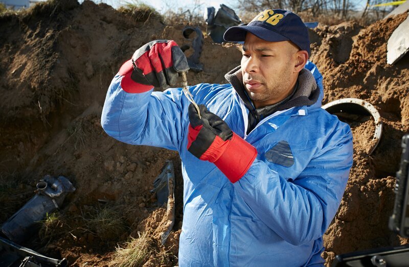 REENACTMENT – An NTSB Investigator (played by a background performer) examines a key piece of wreckage found deep in the impact crater of the plane crash that killed world famous golfer Payne Stewart. (Photo Credit: Cineflix/​Ian Watson) – Bild: National Geographic Channel