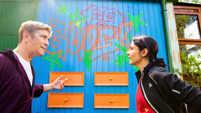 Als Fritz Fuchs (Guido Hammesfahr, l.) mit Schuldirektorin Ina Reuter (Pegah Ferydoni) vom Joggen zurückkommt, hat ein Unbekannter ein Graffiti auf den Bauwagen gesprüht. Ina kommt die Handschrift von ihrer Schule her bekannt vor. – Bild: ZDF und Andrea Hansen Fotografie