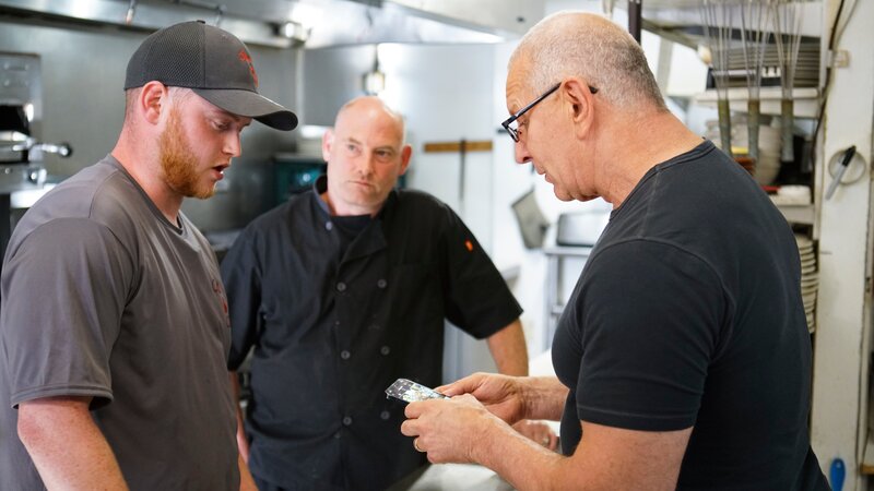 Chef Robert Irvine talks with Blake, son of Mark/​ sous chef at Steak N’ Stuff, in the kitchen of the restaurant. They are joined by Mark Lelushce, owner of Steak N’ Stuff; as seen on Food Network’s Restaurant:Impossible Season 20. – Bild: Discovery, Inc.