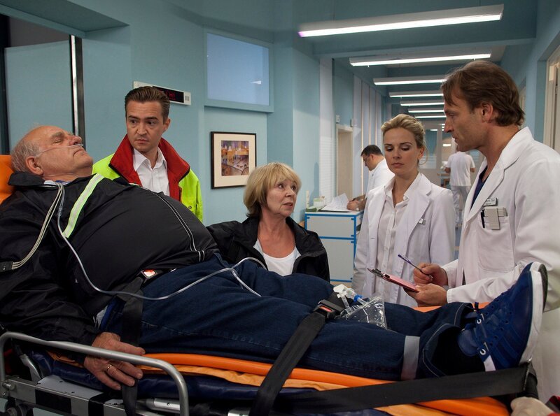 Eduard Runge (Jochen Senf, liegend) ist bei einem Ausflug mit seiner Frau Annerose (Madeleine Lierck-Wien, 2. von links) zusammengebrochen. Dr. Martin Stein (Bernhard Bettermann, rechts) und Dr. Isabel Dahl (Denise Zich, 2. von rechts) nehmen ihn in der Sachsenklinik auf. Notarzt (Komparse, links). – Bild: MDR/​Peter Krajewsky