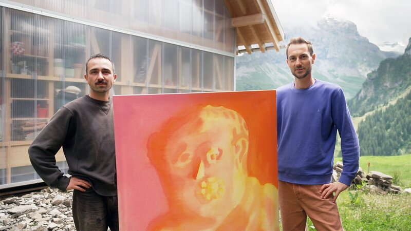 Florian Maritz präsentiert «Kulturplatz»-Host Nino Gadient (rechts) sein gemaltes Kunstwerk auf der Alp Gitschenen vor einer Bergkulisse. – Bild: SRF