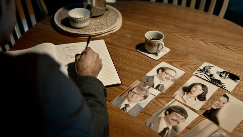 Eine Person macht sich Notizen und schaut sich Fotos auf einem Tisch an. – Bild: A+E NETWORKS