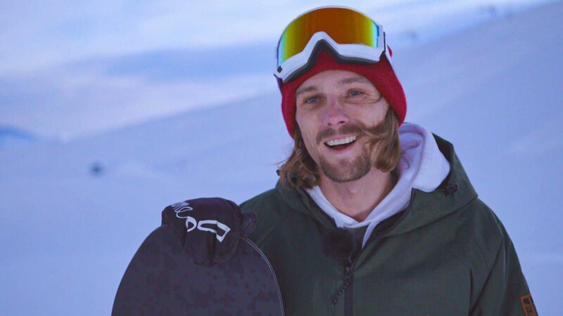 Eine Person steht in einer verschneiten Umgebung und hält ein Snowboard, gekleidet in eine grüne Jacke, einen weißen Hoodie, eine rote Mütze und Skibrille. – Bild: Bergblick