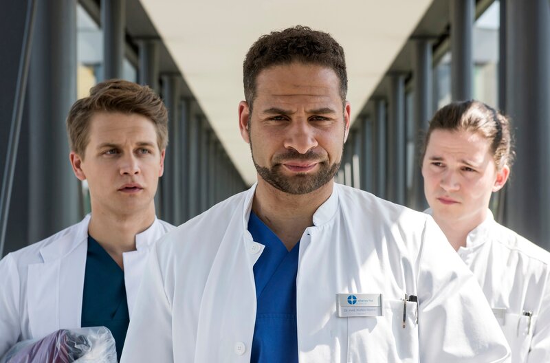 Moreau nimmt sich die beiden Neuen zur Brust und schickt sie zur Patientin Waltraud. Doch warum sollen sie deswegen in den Keller? V.l.n.r. Tom Zondek (Tilman Pörzgen), Dr. Matteo Moreau (Mike Adler), Mikko Rantala (Luan Gummich). – Bild: ARD/​Jens-Ulrich Koch