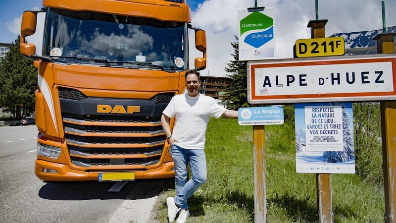 Ep7 – Sebastian in front of the truck (truck on the left of the frame,) left hand leaning on a Alpe d’Huez sign, right thumb on his side, smiling, looking straight to the camera, total – Bild: Warner Bros. Discovery, Inc. or its subsidiaries and affiliates. All rights reserved.