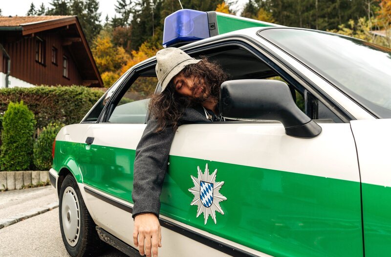 Der Oktoberfest-Tourist Spyros Karamanlis (Nikos Laskaris) ist stark betrunken und wird im Polizeiauto zu seiner Unterkunft gebracht. – Bild: ARD/​Max Bublak/​Max Bublak