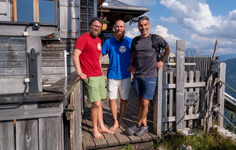 Marcus Fahn (rechts) mit den Hüttenwirten Sebastian Bailey und Andreas Hauber vor der Tegernseer Hütte. – Bild: David Hulik /​ BR, cutflow GmbH /​ BR/​cutflow GmbH/​David Hulik