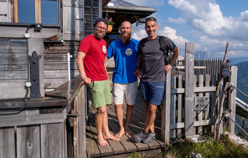 Marcus Fahn (rechts) mit den Hüttenwirten Sebastian Bailey und Andreas Hauber vor der Tegernseer Hütte. – Bild: David Hulik /​ BR, cutflow GmbH /​ BR/​cutflow GmbH/​David Hulik