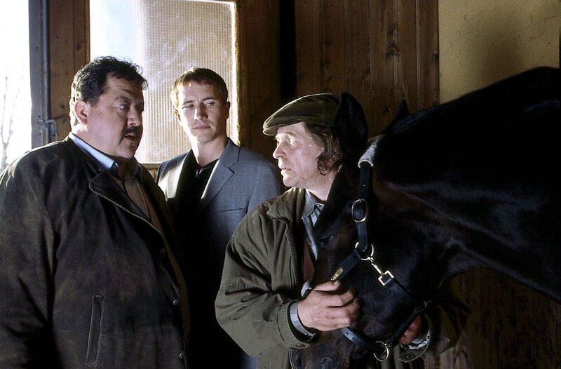 Ein Rennstallbesitzer wurde von seinem Pferd in der Box totgetrampelt, die Box war von aussen verschlossen. Der Stallknecht (Helmut Rühl, re.) scheint den Rosenheim-Cops (Joseph Hannesschläger, li., Markus Böker, mi.) bei ihren Ermittlungen zunächst keine grosse Hilfe zu sein. – Bild: ORF/​ZDF/​Erika Hauri