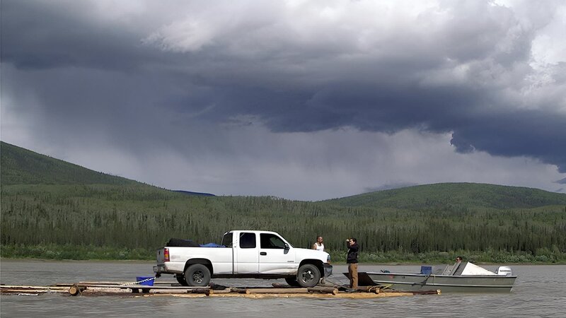 Joey Zuray (R), his sister, Kate Zuray (C), and cousin Zeb (L), transporting a truck down the Yukon River on a hand-built raft during the summer solstice. Joey is sitting, driving the boat. – Bild: For merchandising, publishing & ancillary products, check talent contract, appearance & property releases.