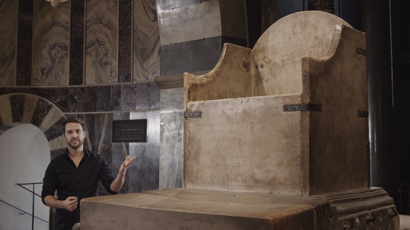 Im Mittelalter sind die Menschen überzeugt, dass Gott persönlich darüber entscheidet, wer zum Herrscher bestimmt ist. Am Thron Karls des Großen in Aachen, neben dem Mirko Drotschmann steht, ist dieser Anspruch bis heute gut abzulesen. – Bild: ZDF und Joachim C. Seck