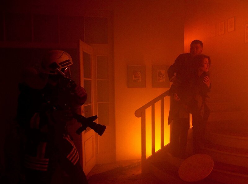 Pia Heilmann (Hendrikje Fitz, rechts) sucht in den Flammen verzweifelt nach ihrem Sohn. Roland Heilmann (Thomas Rühmann) und die Feuerwehrleute versuchen, sie davon abzuhalten. Die Flammen breiten sich immer schneller aus. – Bild: MDR/​Peter Krajewsky