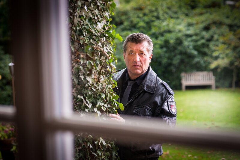 Hans (Bruno F. Apitz) schätzt die Situation ein: Das Ehepaar Baumann sitzt gefesselt in ihrem Wohnzimmer und wird von Gero Broog mit einer Waffe bedroht. – Bild: ZDF/​Boris Laewen