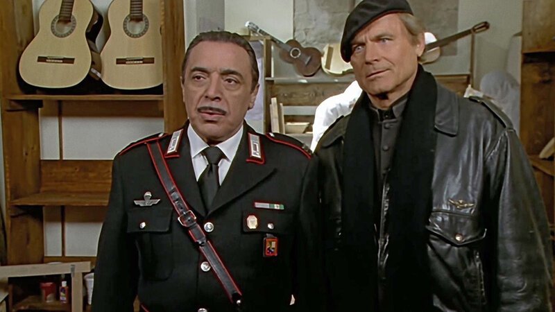 Maresciallo Cecchini und Don Matteo beobachten mit Neugier das Wiedersehen von Capitano Tommasi und seiner alten Freundin Chiara Brini. (Nino Frassica, Terence Hill, v.l.) – Bild: Bibel TV