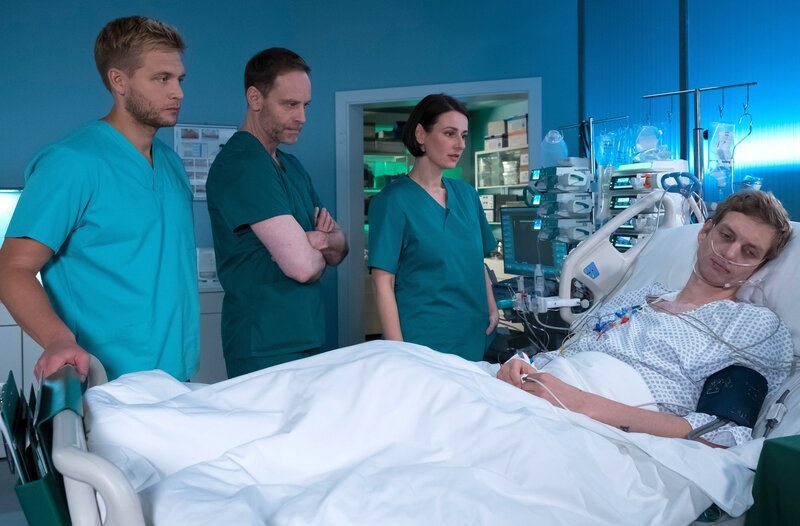 Bea Hölsch (Daniela Wutte, re.) ist mit Fieber umgekippt und hat sich einen Schlüsselbeinbruch zugezogen. Ihren behandelnden Arzt Dr. Martin Stein (Bernhard Bettermann, li.) bringt sie mit ihrem sehr anschmachtenden Verhalten in eine ziemlich unangenehme Situation. Auch Dr. Kathrin Globisch (Andrea Kathrin Loewig, mi.) ist ziemlich irritiert. – Bild: MDR/​Saxonia Media/​Sebastian Kiss