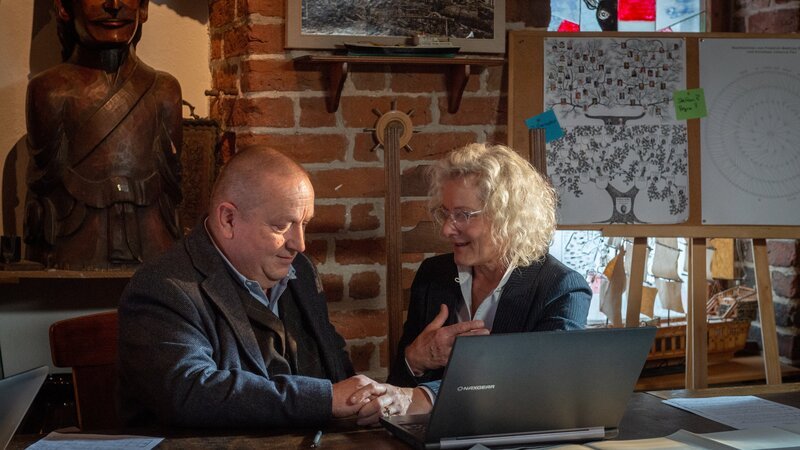 Weil der Tote intensiv Ahnenforschung betrieben hat, hört sich Reuter (Udo Kroschwald, l.) beim Ahnenforscher-Stammtisch um. Dort erfährt er von Charlotte Reuter (Lena Lessing, r.) Erstaunliches: Der Tote hat gerade erst herausgefunden, dass er adoptiert worden ist. Doch warum hat er niemandem davon erzählt, auch seiner Familie nicht? – Bild: Marc Meyerbröker /​ ZDF