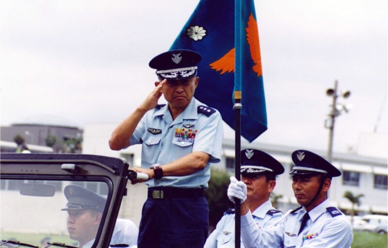 Ein Offizier in einer zeremoniellen Militäruniform salutiert, während er in einem offenen Fahrzeug steht; zwei uniformierte Angehörige halten die Einheitsfahne. – Bild: THE HISTORY CHANNEL /​ A+E Networks /​ Mamoru Sato