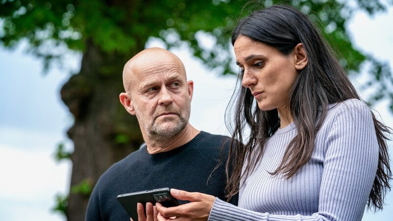 Die Kommissarin schaut gebannt auf ihr Handy, währenddessen sie vom Hauptkommissar beobachtet wird: Jürgen Vogel als Robert Heffler, Aybi Era als Mavi Neumann. – Bild: SRF/​ZDF/​Oliver Feist