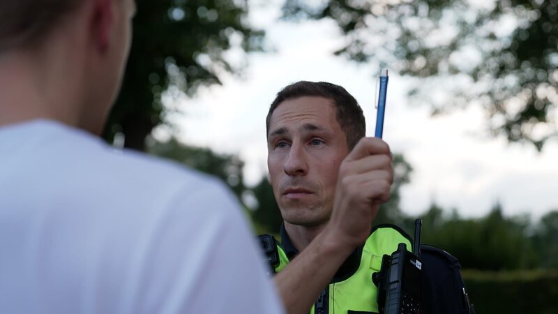 Ein Polizeibeamter führt einen Alkoholtest durch, indem er eine Person auffordert, einem sich bewegenden Objekt mit den Augen zu folgen. – Bild: Warner Bros. Discovery