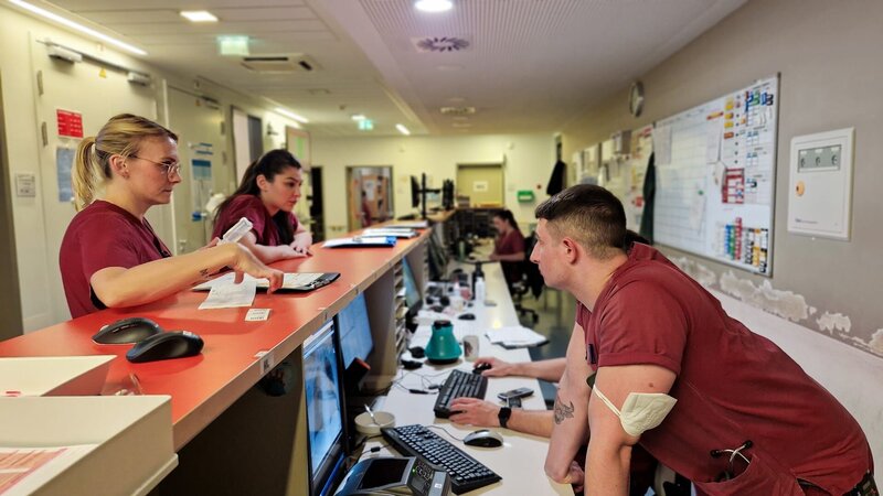 Some members of staff are talking and listening to each other at the orange counter. – Bild: Warner Bros. Discovery, Inc. or its subsidiaries and affiliates