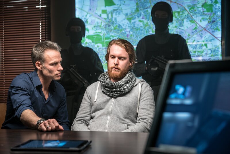 „SOKO Leipzig – Aus der Hölle“: Jan Maybach (Marco Girnth) sitzt mit Bastian Drehwitz (Ole Fischer) im Vernehmungsraum der Kripo. Im Hintergrund sieht man eine an die Wand projizierte Karte und schwer bewaffnete Beamte, sowie im Vordergrund einen Tablet-PC auf dem Tisch und einen Monitor. – Bild: ZDF/​Uwe Frauendorf
