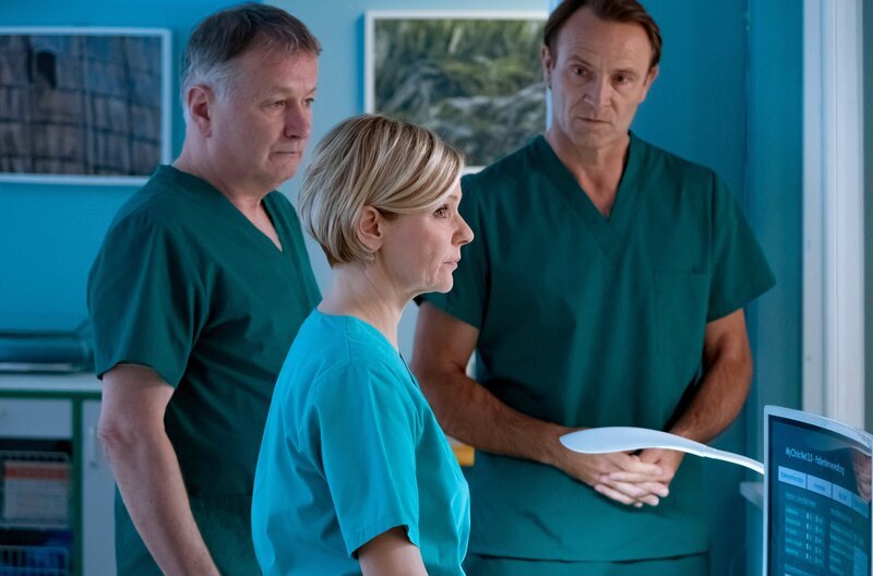 Bea Hölsch (Daniela Wutte, re.) ist mit Fieber umgekippt und hat sich einen Schlüsselbeinbruch zugezogen. Ihren behandelnden Arzt Dr. Martin Stein (Bernhard Bettermann, li.) bringt sie mit ihrem sehr anschmachtenden Verhalten in eine ziemlich unangenehme Situation. Auch Dr. Kathrin Globisch (Andrea Kathrin Loewig, mi.) ist ziemlich irritiert. – Bild: MDR/​Saxonia Media/​Sebastian Kiss