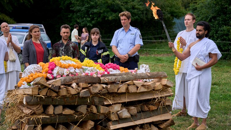Marie (Katharina Wackernagel, 2.v.l.), Mehmet (Cem Ali Gültekin, 3.v.l.), Heino (Sebastian Schwarz, 3.v.r.) und Jenny (Eva Bühnen, M.) verabschieden sich vom verstorbenen Ragulan. Im Hintergrund unterhalten sich Gisbert (Kai Schumann, h.l.) und Barisa (Marta Sroka, h.r.). Prasad (Prashath Lokeswaran, r.) hebt die Fackel, um Ragulans Leichnam zu entzünden (mit Kompars:innen). – Bild: WDR Mediagroup GmbH