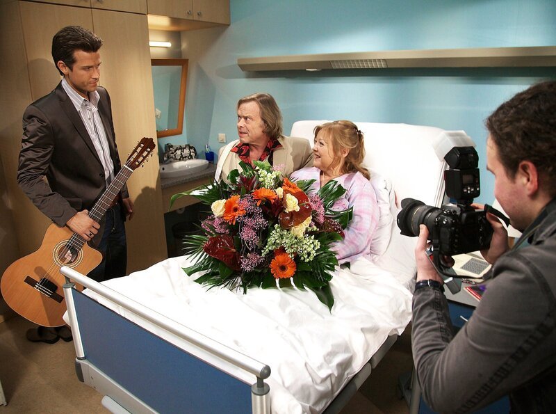 Brigitte Kiesert (Franziska Trögner) ist überrascht, als Tom Peters (Volker Lechtenbrink) mit Manager und Pressefotograf zum Krankenbesuch erscheint. – Bild: MDR/​Wernicke