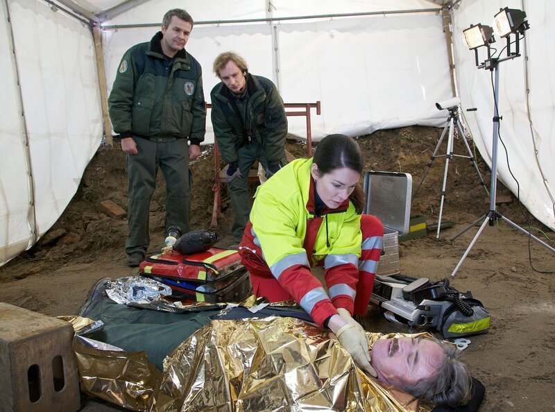 Nachdem Peter Merz (Hartmut Schreier, liegend) die Bombe entschärft hat, bricht er bewusstlos zusammen. Seine Kollegen (Jochen S. Franken, links, Daniel Schlösser, 2. v. von links) bringen Dr. Elena Eichhorn (Cheryl Shepard) zu ihm. Merz hat durch den Aortenriss innere Blutungen und droht zu verbluten. – Bild: MDR/​Wernicke
