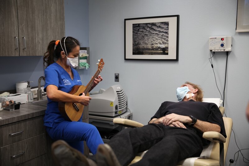 Dr. Lee plays her ukulele for Jerry Harrison post his cheek lipoma removal. – Bild: Circle the Globe Productions