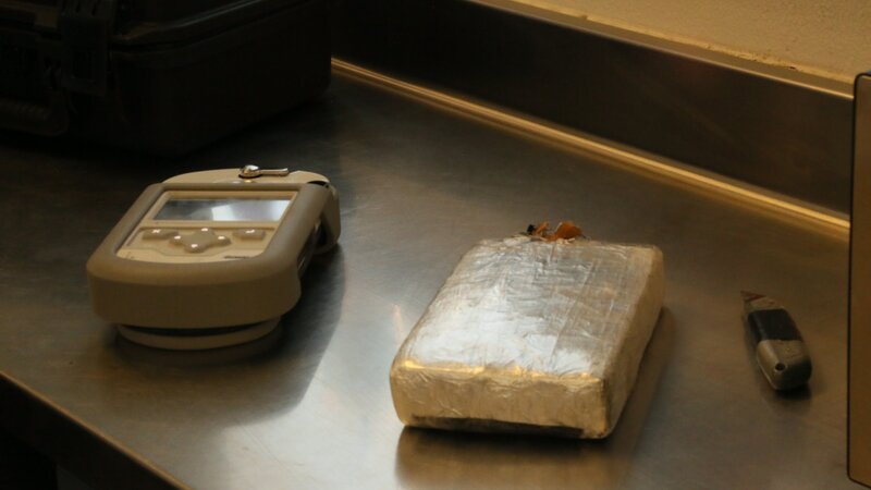 Ein Paket mit mutmaßlichen Betäubungsmitteln, das im Fahrzeug eines Verdächtigen gefunden wurde, liegt auf einem Metalltisch neben einem Gemini-Analysator und einem Teppichmesser und wartet darauf, getestet zu werden. (National Geographic) – Bild: National Geographic /​ National Geographic