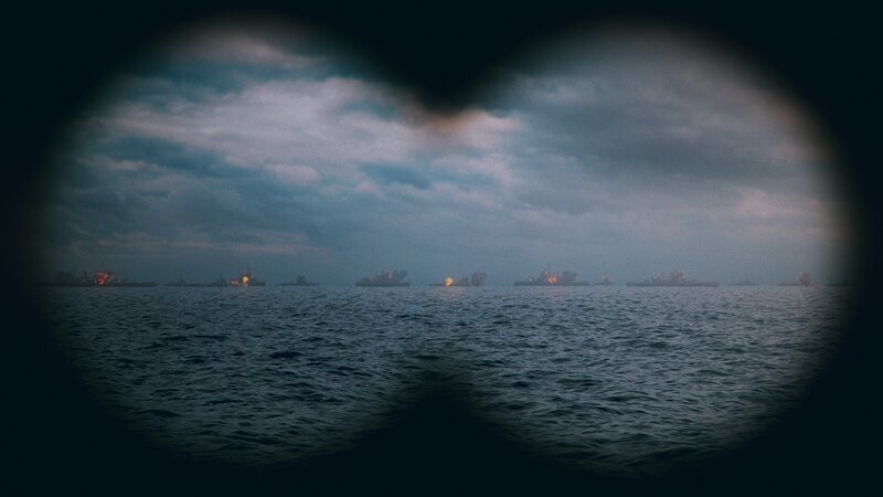 Through binoculars boats in a line on the horizon on fire. – Bild: Disney+