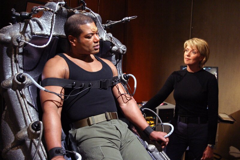 Teal’c (Christopher Judge, li.) soll den Simulator testen. Carter (Amanda Tapping) erklärt ihm genau wie er das Gerät abschalten kann … – Bild: SYFY