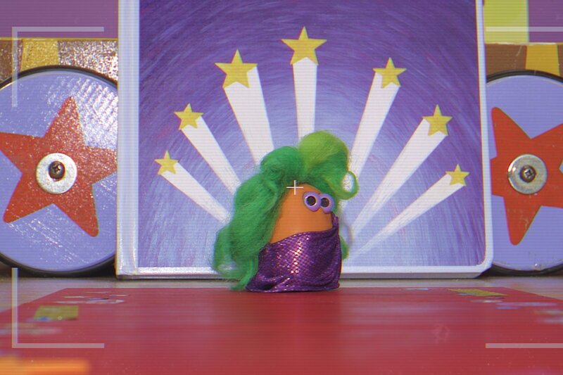 Ein Mini Boing mit grüner Perücke und lilafarbenem Tuch posiert für ein Foto. (Die Mini Boings - Rutschbahn und Fötele, Staffel 2, Folge 7) – Bild: SRF/Aardman Ein Mini Boing mit grüner Perücke und lilafarbenem Tuch posiert für ein Foto. (Die Mini Boings - Rutschbahn und Fötele, Staffel 2, Folge 7) – Bild: SRF/Aardman