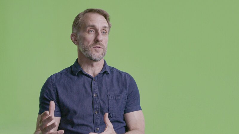 Die Szene zeigt eine Person in einem kurzärmeligen Hemd vor einem einfarbigen grünen Hintergrund. Die Person befindet sich in einer sitzenden Position und gestikuliert mit den Händen, was auf eine Erklärung oder ein Gespräch hindeutet. Die Aufnahme wirkt wie ein Interview oder ein dokumentarisches Gespräch. – Bild: A+E Global Media, THE HISTORY CHANNEL